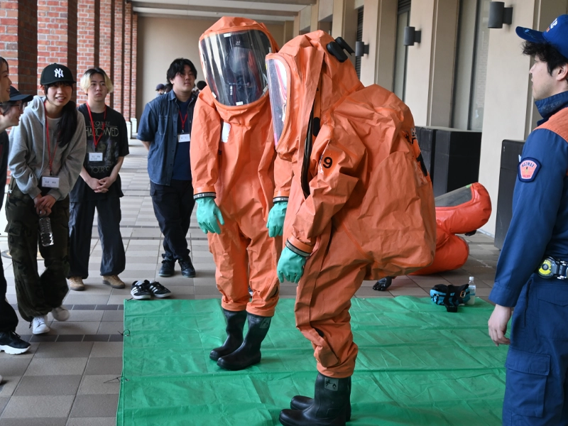 宇宙服のような防災服を着た学生2人は重さを実感