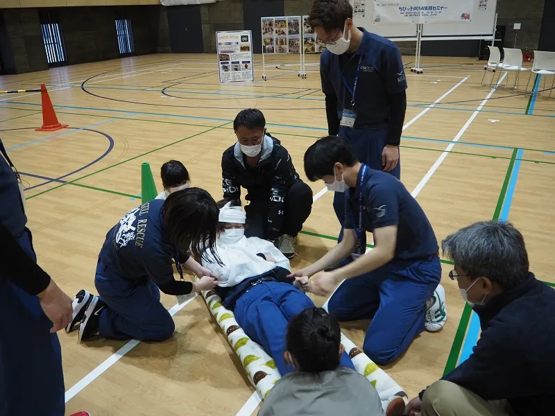 負傷役の学生にケガの手当を行い全員で力を合わせて簡易担架で運びました