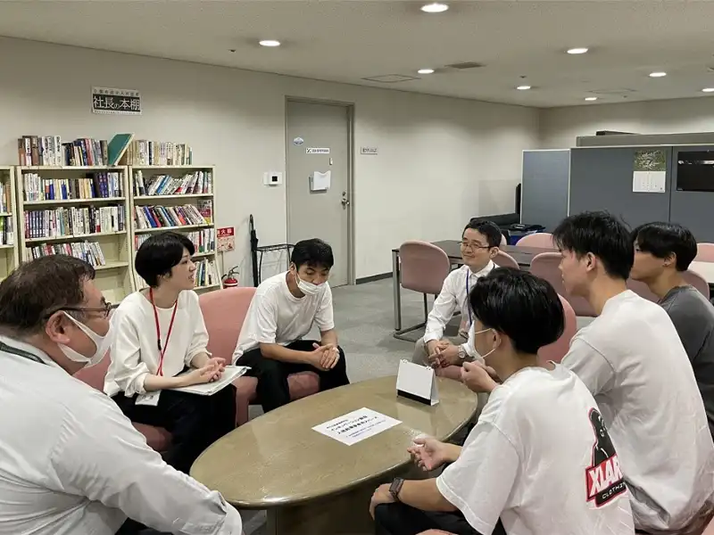 企業育成室の共有スペースで産振財団の皆さんから話を聞かせてもらう学生たち。背景に見えるのは経営に役立つ本を集めた「社長の本棚」
